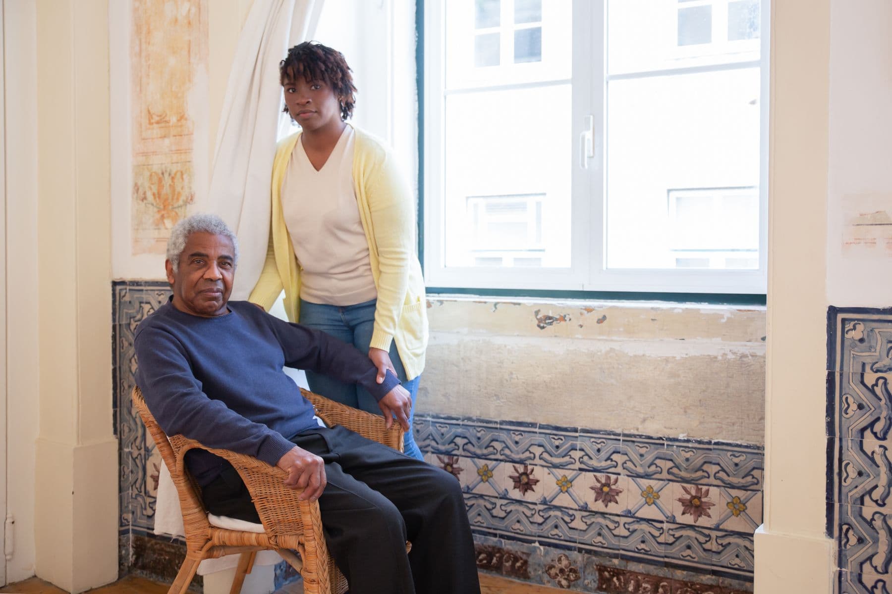 A caregiver in a white shirt stands beside an older man seated by a window at home.