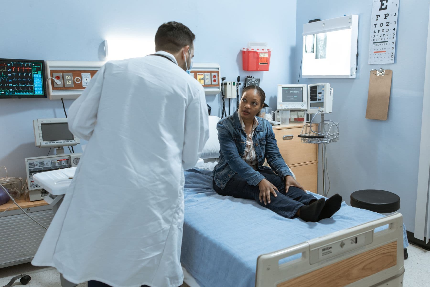 A doctor in a white coat sits with a patient on a bed in a bright room.